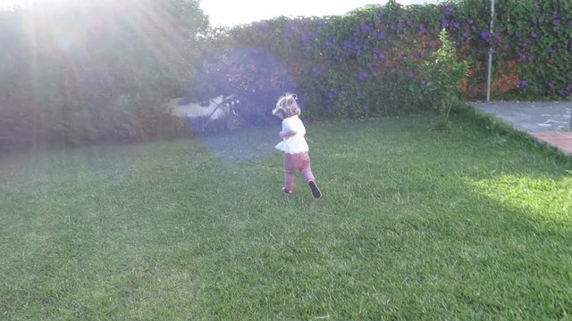 slow motion back two years age blonde baby girl with white shirt running in exterior garden green grass and plants
