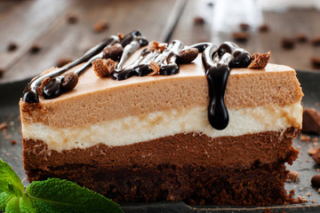 piece of layered cake with chocolate topping close-up on black plate, blurred wooden background