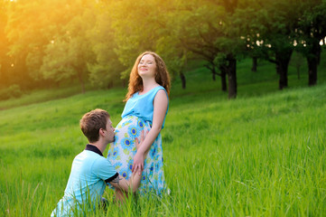 Fototapeta premium Happy pregnant couple standing on meadow at sunset. The guy kisses the girl belly. The girl laughs with delight. Portrait. Horizontal orientation