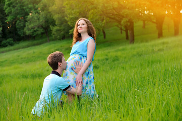 Fototapeta premium Happy pregnant couple playing in the Park at sunset. The guy kisses the girl belly. Portrait. Horizontal orientation