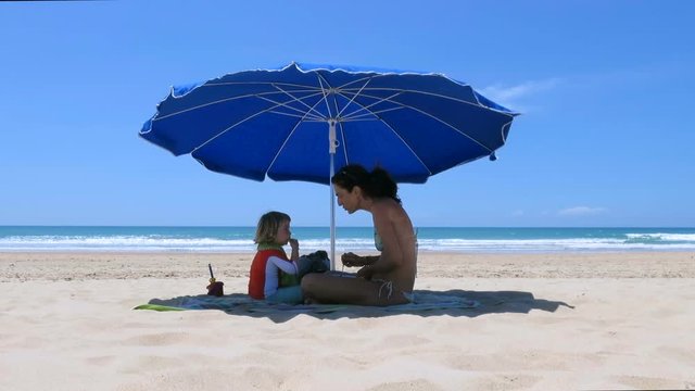 little baby and mother sitting down of parasol at beach eating and sharing food
