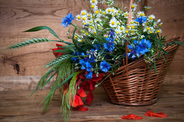 Cornflowers, chamomiles wheat and poppies in basket, wooden back