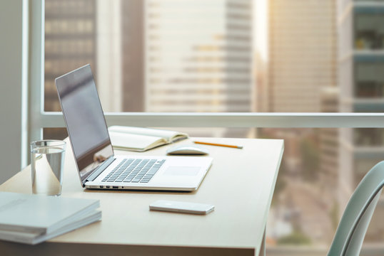 Wooden desk with modern laptop, glass of water, modern cell phone, notebook. Desktop with a view of downtown, Sunrise, Shallow DOF.
