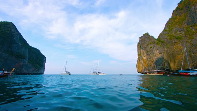 Video UHD - Several private sailboats, bobbing peacefully in the gentle, tropical sea, beneath the towering seacliffs of Phi-Phi Island in southern Thailand.