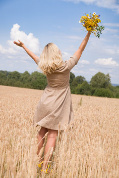 Back view on happy woman with flowers running on the wheat field and enjoying sun. Summer and blue sky background. Girl walking on the meadow. Lifestyle and happiness, freedom concept
