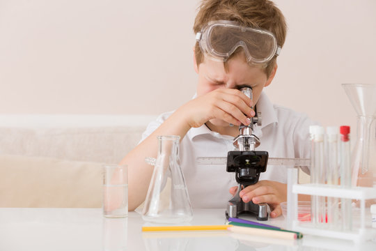 Cute elementary schoolboy looking into microscope at his desk at home. Young scientist making experiments in his home laboratory. Indoors. Child and science. Education concept.