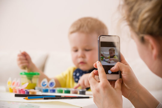 Young mother taking photo of cute little toddler boy in yellow shirt sitting at table and drawing with colorful paints. Early learning. Creative. Toddler drawing. Mom making picture on mobile phone