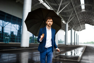 Picture of  young  businessman holding  suitcase and umbrella standing at rainy station looking in camera