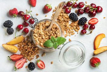 Healthy breakfast ingredients. Oat granola in jar served with slices of peach, strawberry, sweet cherries and blackberries on light marble background, top view, horizontal composition