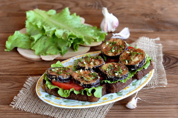 Dark bread toasts with grilled eggplant, fresh tomatoes, lettuce, garlic, dill. Green lettuce on the blackboard, garlic. Sandwiches with vegetables on plate on wooden background