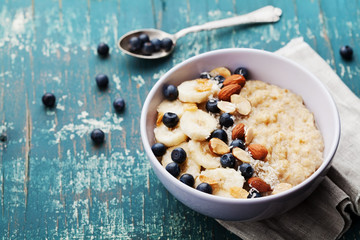 Bowl of fresh oatmeal porridge with banana, blueberries, almonds, coconut and caramel sauce on teal rustic table, hot and healthy food for Breakfast