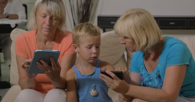 Grandmothers and grandson spending time at home. One grandma using tablet computer and another playing with kid on mobile phone