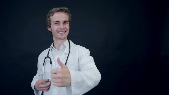Smiling Doctor showing thumbs up, standing on a black background putting on a a stethoscope. 1080p.