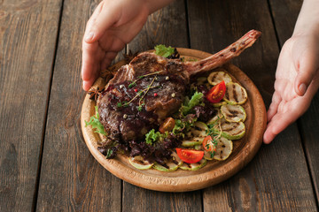 Portion of roasted rib steak on board serving. Hands put on wooden table dish with grilled pork on bone