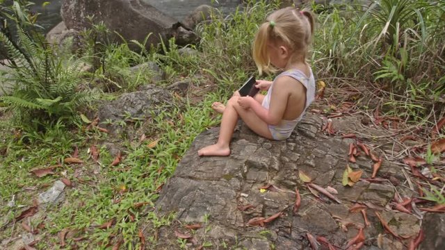 Little Girl Plays with Phone on Stone against Waterfall