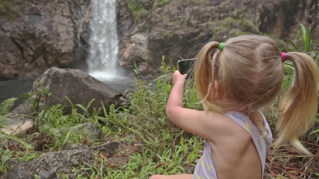 Closeup Little Girl Plays with Phone on Stone against Waterfall