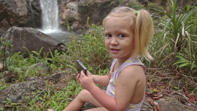 Closeup Little Girl Plays with Phone on Stone against Waterfall