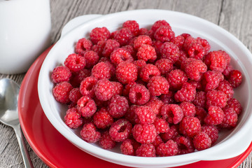  Fresh raspberries.  White porcelain bowl with fresh raspberries on a red plate on a wooden table.