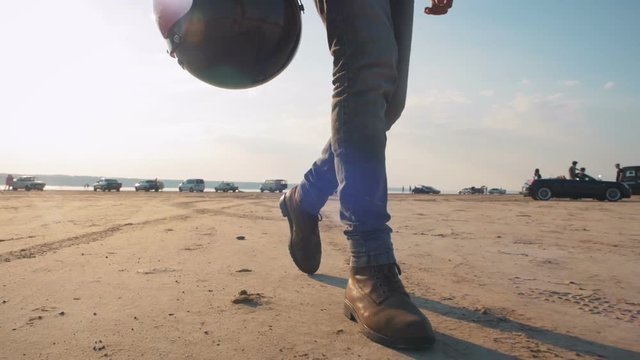 Close up shot of motorcyclist walking in desert with his helmet during sunset