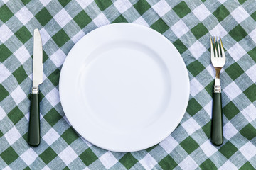 Knife, white plate and fork on green picnic tablecloth. Flat lay