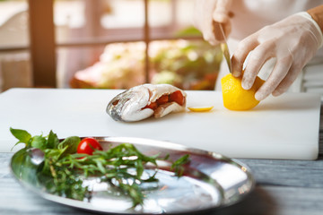 Hand with knife cuts lemon. Raw fish stuffed with tomatoes. Fresh lemon for seafood dish. Chef's new recipe.