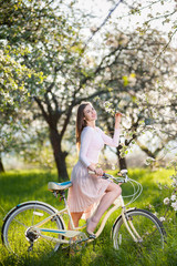 Pretty female with a retro bicycle in the lush green grass enjoying flowers of a blossoming tree in the ray of sunshine in a spring garden