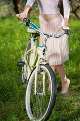 Fototapeta na wymiar Graceful female dressed in a light skirt and top keeps retro bike against the background of fresh greenery in spring. Close-up