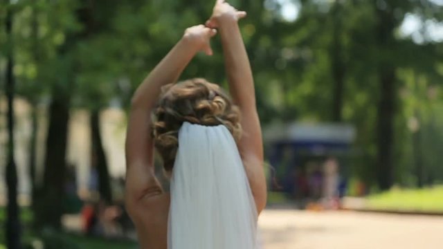 Cheerful bride in exellent white dress with long veil playfully lifting her hands when walking by sunny alley in the park