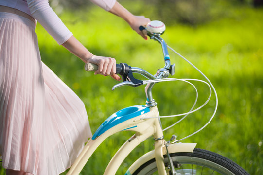 Hands of a girl dressed in a light skirt holding handbrake on vintage bicycle on the blurred background of fresh greenery. Close-up