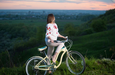 Young female in Ukrainian embroidery holding vintage bicycle at hill and looking into the distance, on a blurred background of greenery and small town at sunset