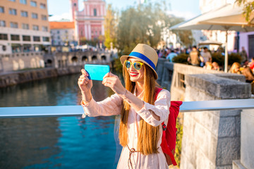 Fototapeta na wymiar Young female traveler making selfie photo with phone on the bridge in the center of Ljubljana city in Slovenia. Traveling Slovenia