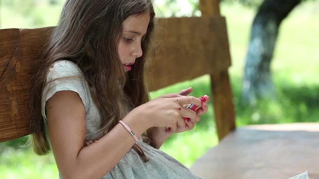 Teen girl sits on the bench in garden and uses red smartphone. Beautiful girl with smartphone sits on the bench. Girl playing the game on his smartphone. Female with smartphone