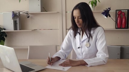 Happy indian therapist wearing in white lab coat sitting at the wooden table in office. Mixed race professional female work as physician in hospital fill recipe. Multicultural model smiling kindly on