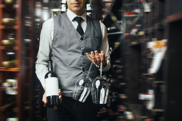 Close up of arms of skillful male sommelier standing in liquor store. He is holding a bottle of wine and two glasses. The man is smiling. Copy space in right side - obrazy, fototapety, plakaty