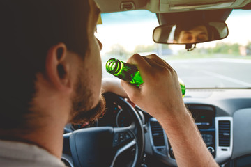 Drunk young man driving a car with a bottle of beer. Don't drink and drive concept. Driving under...