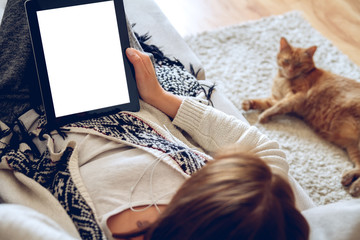 Woman in home cozy clothes lying on a sofa using tablet with headphones, looking at a lazy red cat sprawled on the carpet beside. Online education concept. e-learning. back view. Pet shop concept - Powered by Adobe