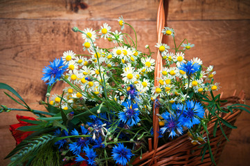Cornflowers, chamomiles wheat and poppies in basket, wooden back