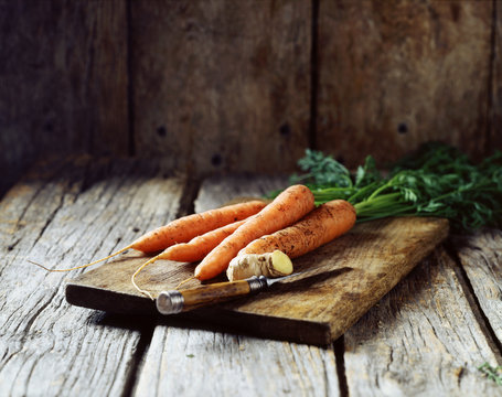 Fototapeta Organic muddy carrots and carrot tops, ginger with skin on, rustic wooden board on wooden table, vintage knife