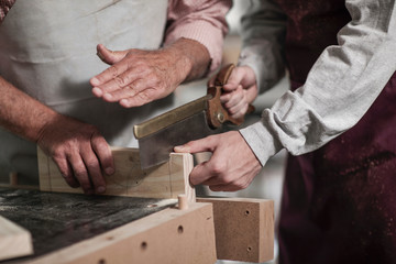 Cropped shot of senior carpenter demonstrating sawing to trainee in workshop