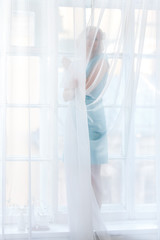 Young blond woman standing behind transparent white curtains