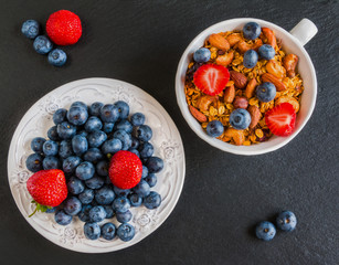 Breakfast bowl with granola made from oat flakes, dried fruits and nuts, and fresh blueberries and strawberries. Black stone background, top view.