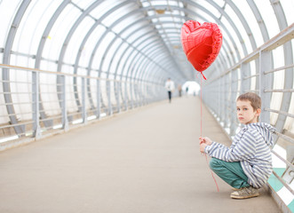 boy with red heart sitting on the sidelines. sad child sits on his haunches and holding a red...