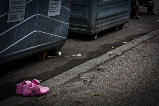 Scarpette rosa da bambina abbandonate su un marciapiede accanto a dei cassonetti della spazzatura.
