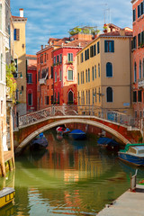 Fototapeta premium Colorful narrow lateral canal and pedestrian bridge in Venice with docked boats, Italy