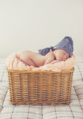 Adorable little guy,in a gray knitted cap,tucked under his legs and put the handle under his head,sound asleep on the soft beige blanket in a large wicker basket high,sweet sleeping newborn baby