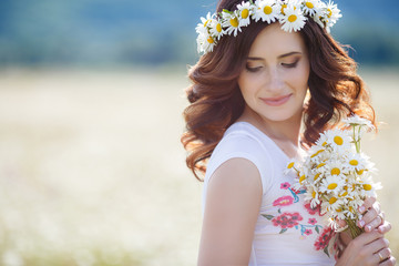 Cute pregnant brunette woman in white t-shirt with a bouquet of white daisies on his head wears a wreath of white flowers,chamomile,summer,walking alone in a beautiful field of blooming white daisies