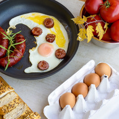 Breakfast - sausages and eggs frying in a pan and served with bread, tomatoes, egg shell on a grey stone board
