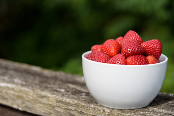 Fresh strawberries in a white bowl, on a deck railing with greenery in the background
