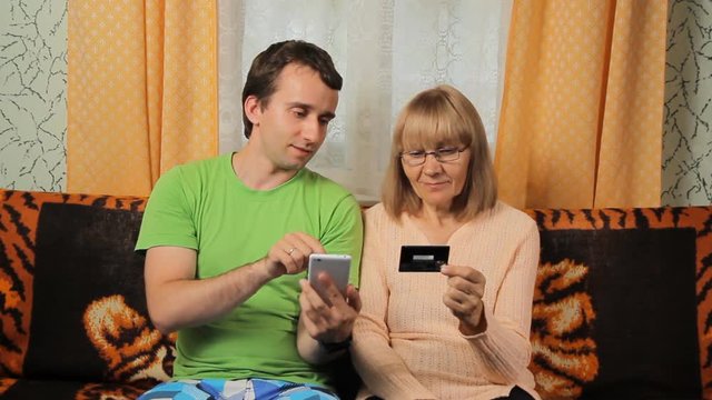Adult son and elderly mother making online banking with credit card by phone. Mother and son smiling at home on sofa