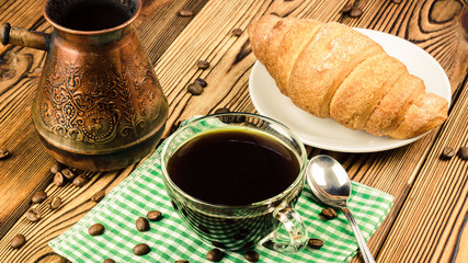 Cup of black coffee on green napkin with croissant, turkish coffee pot, wooden table in cafe